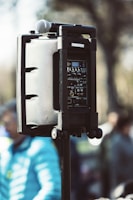 A portable speaker system is mounted on a stand, featuring a microphone placed on top. The speaker system's control panel is visible, showing various dials and switches. The image is taken outdoors, with a softly blurred background of people and trees, suggesting a public outdoor event.