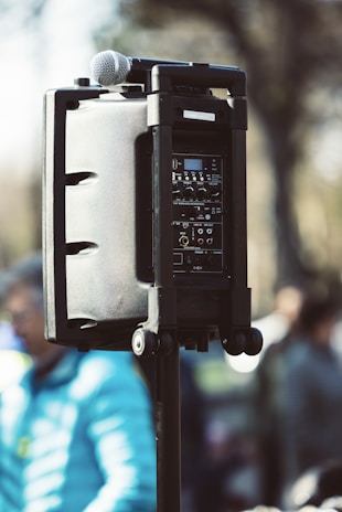 A portable speaker system is mounted on a stand, featuring a microphone placed on top. The speaker system's control panel is visible, showing various dials and switches. The image is taken outdoors, with a softly blurred background of people and trees, suggesting a public outdoor event.