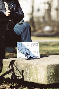 A person is sitting outdoors on a stone platform, wearing a dark coat and jeans. They are holding a sign that reads, 'Be a part of the solution not the pollution.' The image is set in a natural environment with blurred trees in the background, suggesting a park or open space.