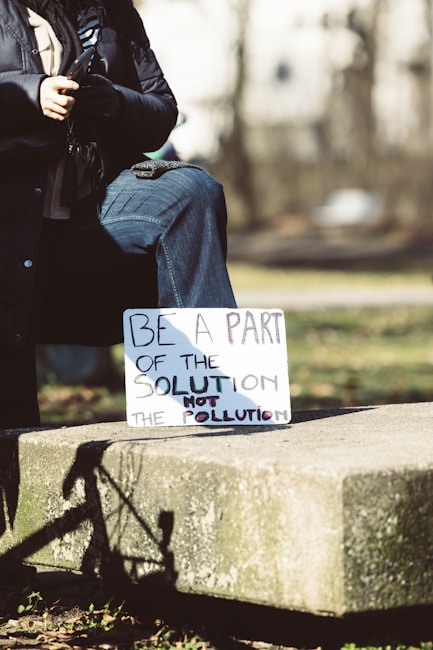 A person is sitting outdoors on a stone platform, wearing a dark coat and jeans. They are holding a sign that reads, 'Be a part of the solution not the pollution.' The image is set in a natural environment with blurred trees in the background, suggesting a park or open space.