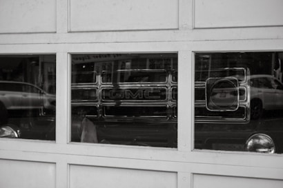 A reflection of a GMC vehicle is visible in the window panes of a garage door. The image is in black and white, with reflections of other parked cars on the street also appearing in the glass.