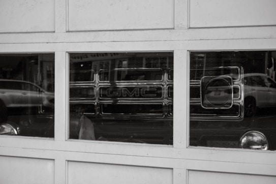 A reflection of a GMC vehicle is visible in the window panes of a garage door. The image is in black and white, with reflections of other parked cars on the street also appearing in the glass.