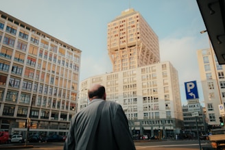 A man dressed in a suit stands at a city intersection, gazing up at a modern high-rise building. Surrounding him are other tall structures, including distinctive architectural designs. Traffic signs and a few vehicles are visible, suggesting an urban environment.