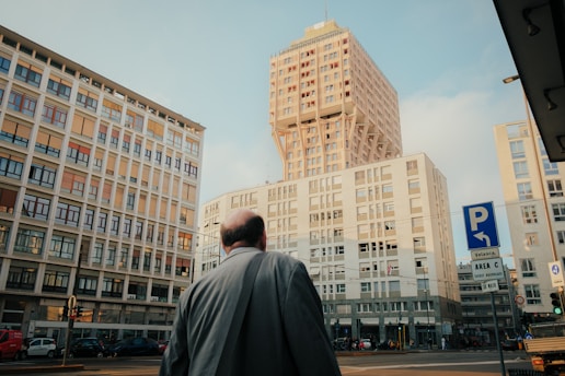 A man dressed in a suit stands at a city intersection, gazing up at a modern high-rise building. Surrounding him are other tall structures, including distinctive architectural designs. Traffic signs and a few vehicles are visible, suggesting an urban environment.