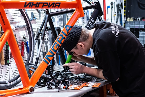 A person is working on assembling or repairing a bicycle in a workshop. The individual is focused on a component, using tools to work on the frame. The workshop is equipped with various tools hanging on a pegboard and other bicycle parts are visible around the workspace.