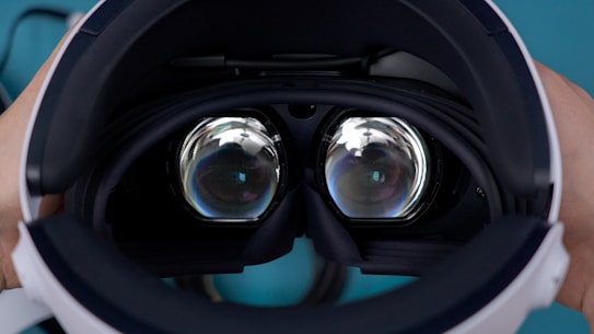 A pair of hands is holding a virtual reality headset, showing the dual lenses inside. The lenses are large and have a reflective surface, capturing light and reflections from the surroundings. The design and close-up view focus on the technology and optics of the device.
