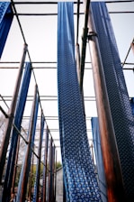 Long strips of blue fabric with white circular patterns hang vertically from a wooden structure. The setup appears to be part of a traditional fabric drying or dyeing process under the open sky. The wooden beams are arranged in a grid pattern, and the fabric gently sways in the wind.