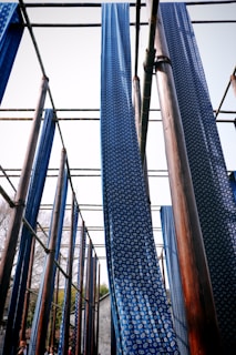Long strips of blue fabric with white circular patterns hang vertically from a wooden structure. The setup appears to be part of a traditional fabric drying or dyeing process under the open sky. The wooden beams are arranged in a grid pattern, and the fabric gently sways in the wind.