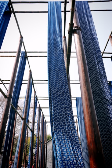 Long strips of blue fabric with white circular patterns hang vertically from a wooden structure. The setup appears to be part of a traditional fabric drying or dyeing process under the open sky. The wooden beams are arranged in a grid pattern, and the fabric gently sways in the wind.