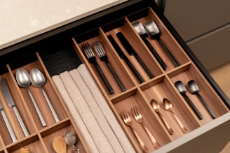 A neatly organized wooden cutlery drawer contains various types of utensils, including stainless steel and black-handled knives, forks, and spoons. Cloth napkins are folded and stored in one section, with silver and copper-toned utensils placed separately.