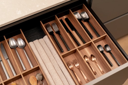 A neatly organized wooden cutlery drawer contains various types of utensils, including stainless steel and black-handled knives, forks, and spoons. Cloth napkins are folded and stored in one section, with silver and copper-toned utensils placed separately.
