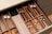A neatly organized wooden cutlery drawer contains various types of utensils, including stainless steel and black-handled knives, forks, and spoons. Cloth napkins are folded and stored in one section, with silver and copper-toned utensils placed separately.