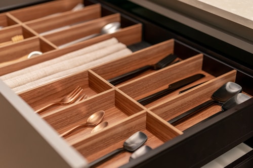 An organized kitchen drawer contains various utensils neatly arranged in wooden dividers. The utensils include spoons, forks, knives, and napkins. The dividers provide separate compartments for each type of cutlery, enhancing the aesthetic of the drawer's organization.