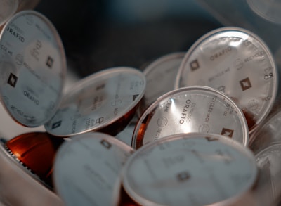 A close-up view of coffee capsules with shiny foil lids labeled 'Espresso Orafio' and containing various recycling symbols. The capsules are stacked and overlapping each other, surrounded by a soft lighting that highlights their metallic texture.
