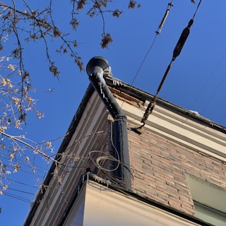 A corner of a brick building is seen with a drainpipe extending from the roof, partially covered with icicles. Surrounding the pipe are bare tree branches and overhead cables. The sky is clear and blue.