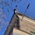 A corner of a brick building is seen with a drainpipe extending from the roof, partially covered with icicles. Surrounding the pipe are bare tree branches and overhead cables. The sky is clear and blue.