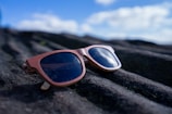 A pair of stylish sunglasses with wooden frames is placed on a textured, dark rock surface. The background features a bright blue sky with a few fluffy clouds, slightly out of focus.