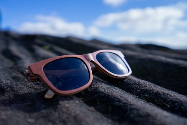 A pair of stylish sunglasses with wooden frames is placed on a textured, dark rock surface. The background features a bright blue sky with a few fluffy clouds, slightly out of focus.