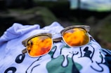 A pair of sunglasses with reflective orange lenses lies atop a white piece of clothing featuring abstract designs in black and green. The reflection in the lenses shows a scenic view with clouds.