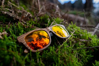A pair of wooden-frame sunglasses with vibrant orange reflective lenses rests on lush, green moss. The reflection in the lenses suggests an outdoor setting, possibly reflecting trees or a forest canopy, creating a natural and organic feel.
