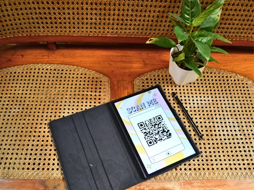 A wooden and woven cane chair is visible with a tablet displaying a QR code labeled 'scan me'. Next to it is a stylus and a small potted plant with glossy green leaves in a white pot.