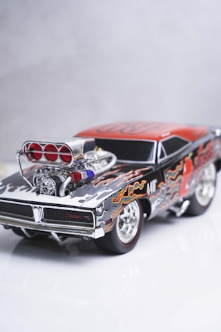 A detailed model of a classic muscle car featuring a black and red color scheme with flame decals. The car has a prominent, polished engine with visible air intakes and chrome detailing. The wheels are styled with chrome rims, enhancing the overall aesthetic of power and speed.
