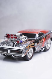 A detailed model of a classic muscle car featuring a black and red color scheme with flame decals. The car has a prominent, polished engine with visible air intakes and chrome detailing. The wheels are styled with chrome rims, enhancing the overall aesthetic of power and speed.