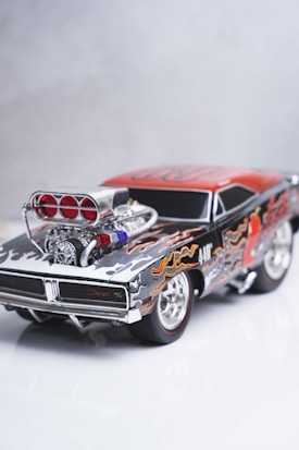 A detailed model of a classic muscle car featuring a black and red color scheme with flame decals. The car has a prominent, polished engine with visible air intakes and chrome detailing. The wheels are styled with chrome rims, enhancing the overall aesthetic of power and speed.
