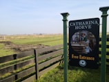 A scenic landscape featuring a grassy field with a wooden fence running through it. In the foreground, there is a sign reading 'CATHARINA HOEVE Free Entrance', with an illustration of a historical scene involving a woman in traditional attire and a child. The background shows a wide expanse of flat land with distant trees and a faint view of a windmill under a clear blue sky.
