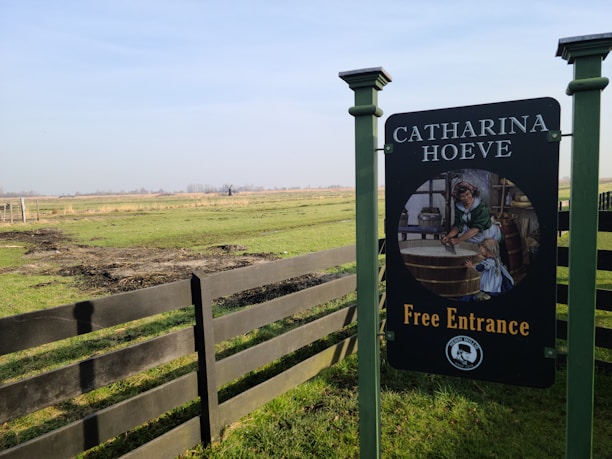A scenic landscape featuring a grassy field with a wooden fence running through it. In the foreground, there is a sign reading 'CATHARINA HOEVE Free Entrance', with an illustration of a historical scene involving a woman in traditional attire and a child. The background shows a wide expanse of flat land with distant trees and a faint view of a windmill under a clear blue sky.