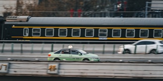 A green taxi is driving on a road parallel to a moving train in the background. The train has a black and yellow color scheme, and the scene captures a dynamic urban setting. Another car is visible slightly blurred in the background, indicating motion.