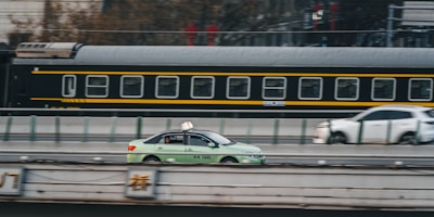 A green taxi is driving on a road parallel to a moving train in the background. The train has a black and yellow color scheme, and the scene captures a dynamic urban setting. Another car is visible slightly blurred in the background, indicating motion.