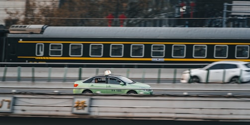 A green taxi is driving on a road parallel to a moving train in the background. The train has a black and yellow color scheme, and the scene captures a dynamic urban setting. Another car is visible slightly blurred in the background, indicating motion.