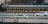 A green taxi is driving on a road parallel to a moving train in the background. The train has a black and yellow color scheme, and the scene captures a dynamic urban setting. Another car is visible slightly blurred in the background, indicating motion.