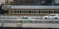 A green taxi is driving on a road parallel to a moving train in the background. The train has a black and yellow color scheme, and the scene captures a dynamic urban setting. Another car is visible slightly blurred in the background, indicating motion.