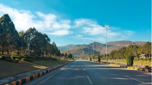 A wide, carpeted road lined with young trees and modern streetlights under a clear blue sky in Faisal Town 2