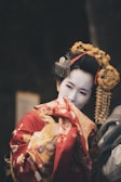 A woman wearing traditional Japanese attire is dressed in a vibrant red and gold kimono adorned with floral patterns. Her face is painted white, and she has an elaborate hairstyle featuring intricate hair ornaments and bright yellow decorations.