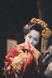 A woman wearing traditional Japanese attire is dressed in a vibrant red and gold kimono adorned with floral patterns. Her face is painted white, and she has an elaborate hairstyle featuring intricate hair ornaments and bright yellow decorations.
