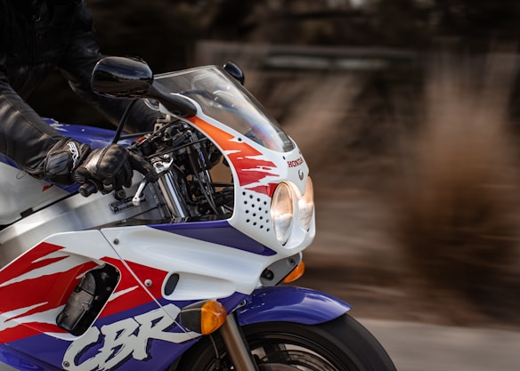 A close-up of a Honda motorcycle with a colorful design in red, white, and blue. The rider is wearing black leather gloves and is gripping the handlebars. The background is blurred, suggesting motion.
