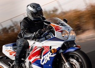 A motorcyclist in full gear, including a helmet and leather jacket, is riding a sport motorcycle on a road. The motorcycle is predominantly white with blue, red, and yellow accents, with a prominent CBR logo on its side. The background is blurred, indicating high speed or motion.