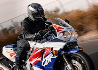 A motorcyclist in full gear, including a helmet and leather jacket, is riding a sport motorcycle on a road. The motorcycle is predominantly white with blue, red, and yellow accents, with a prominent CBR logo on its side. The background is blurred, indicating high speed or motion.