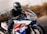 A motorcyclist in full gear, including a helmet and leather jacket, is riding a sport motorcycle on a road. The motorcycle is predominantly white with blue, red, and yellow accents, with a prominent CBR logo on its side. The background is blurred, indicating high speed or motion.