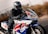 A motorcyclist in full gear, including a helmet and leather jacket, is riding a sport motorcycle on a road. The motorcycle is predominantly white with blue, red, and yellow accents, with a prominent CBR logo on its side. The background is blurred, indicating high speed or motion.