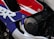 Close-up view of a motorcycle fairing with a vibrant design featuring red, blue, and white colors. The word 'Honda' is engraved on a circular, metallic engine cover. The area includes bolts and part of the chassis, highlighting motorcycle components and branding.