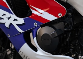 Close-up view of a motorcycle fairing with a vibrant design featuring red, blue, and white colors. The word 'Honda' is engraved on a circular, metallic engine cover. The area includes bolts and part of the chassis, highlighting motorcycle components and branding.