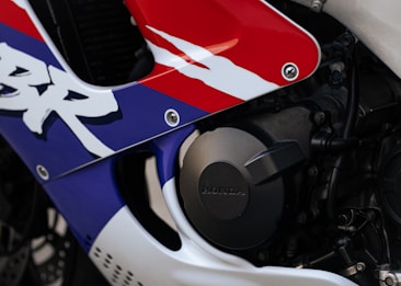 Close-up view of a motorcycle fairing with a vibrant design featuring red, blue, and white colors. The word 'Honda' is engraved on a circular, metallic engine cover. The area includes bolts and part of the chassis, highlighting motorcycle components and branding.