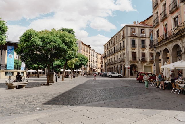 A charming European town square featuring historic buildings with balconies and large archways. Trees provide shade, and there are outdoor seating areas with tables and umbrellas. People are strolling, interacting, and sitting on a bench, giving the area a lively but relaxed atmosphere. Cars are parked along the cobblestone and paved street.