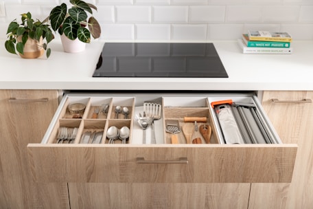A modern kitchen scene featuring an open drawer filled with organized kitchen utensils and cutlery, including forks, spoons, knives, and various cooking tools. The countertop above the drawer holds a potted plant and a couple of stacked books on one side, with a sleek black stove embedded in the white countertop. The kitchen cabinets are made of light wood and there's a white tiled backsplash creating a clean and contemporary look.