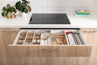 A modern kitchen scene featuring an open drawer filled with organized kitchen utensils and cutlery, including forks, spoons, knives, and various cooking tools. The countertop above the drawer holds a potted plant and a couple of stacked books on one side, with a sleek black stove embedded in the white countertop. The kitchen cabinets are made of light wood and there's a white tiled backsplash creating a clean and contemporary look.