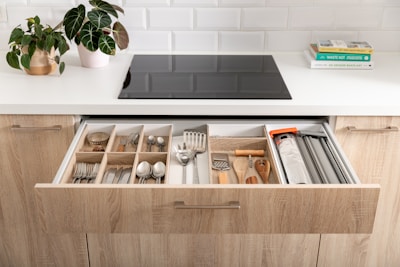 A modern kitchen scene featuring an open drawer filled with organized kitchen utensils and cutlery, including forks, spoons, knives, and various cooking tools. The countertop above the drawer holds a potted plant and a couple of stacked books on one side, with a sleek black stove embedded in the white countertop. The kitchen cabinets are made of light wood and there's a white tiled backsplash creating a clean and contemporary look.